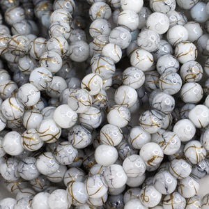 May include: A collection of white marble-like beads with gold and gray veining. The beads are round and have a glossy finish. These beads are suitable for jewelry making, crafting, and other creative projects.