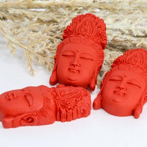 May include: Three red Buddha head sculptures with closed eyes and ornate headdresses. The sculptures are arranged on a white surface with dried plants in the background. The heads are carved with detailed facial features.