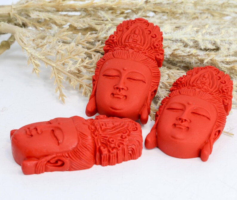 May include: Three red Buddha head sculptures with closed eyes and ornate headdresses. The sculptures are arranged on a white surface with dried plants in the background. The heads are carved with detailed facial features.