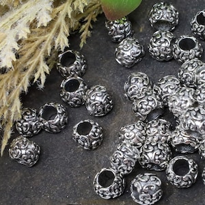 May include: A collection of silver-colored, ornate beads. Each bead is round with a hollow center and features a detailed, swirling pattern. The beads are scattered on a dark, textured surface, with some dried foliage in the background.