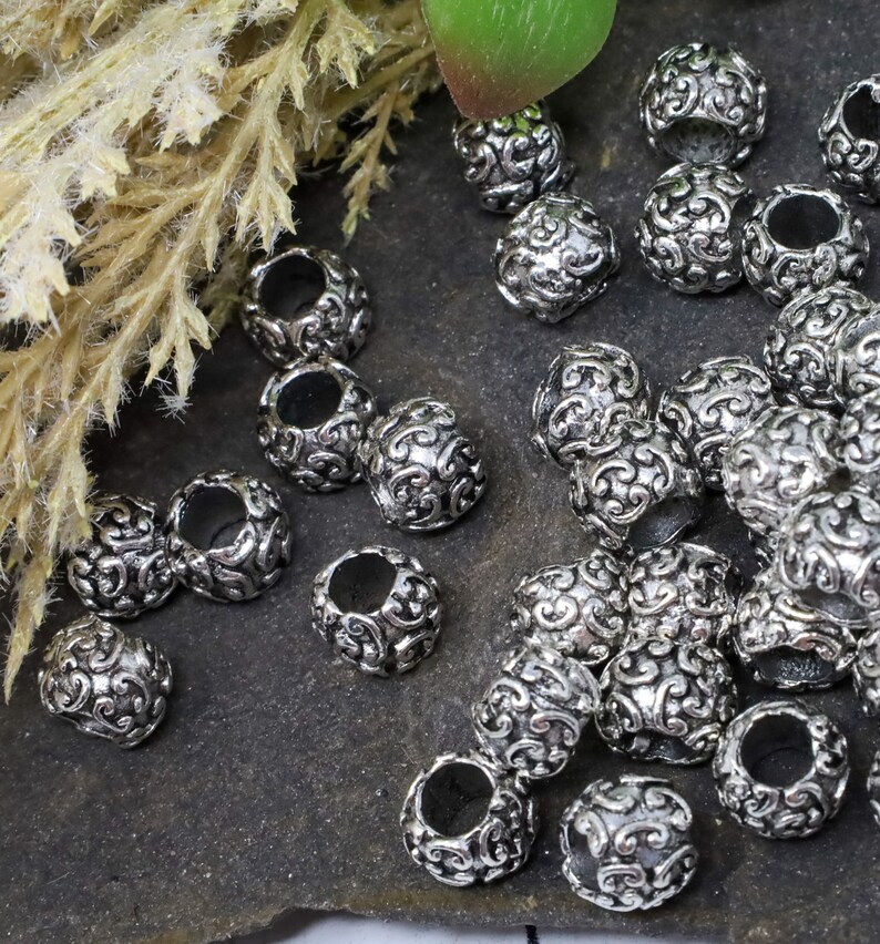 May include: A collection of silver-colored, ornate beads. Each bead is round with a hollow center and features a detailed, swirling pattern. The beads are scattered on a dark, textured surface, with some dried foliage in the background.