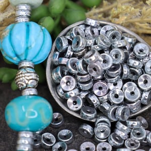May include: A close-up of a bowl filled with silver metal beads. The beads are round with a hole in the center. The bowl is sitting on a surface with a turquoise and silver beaded necklace.