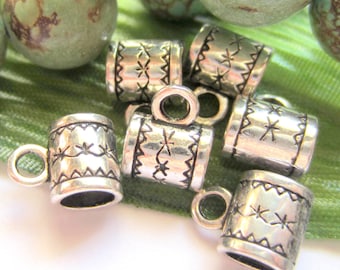 May include: Close-up of several small, silver-colored metal beads shaped like miniature mugs. Each bead has a handle and is decorated with etched lines and star-like patterns. The beads are scattered on a green surface.