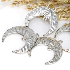 May include: Close-up of several silver-colored crescent moon charms. Each charm has a textured surface and a small loop for attaching to jewelry. The charms are arranged on a white surface, with some dried plant stems in the background.