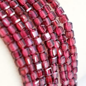 May include: Close-up of strands of faceted, deep red gemstone beads. The beads are square-cut and strung together, creating a rich, textured appearance. The color is a vibrant, saturated red, with hints of light reflecting off the facets.