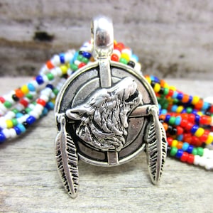 May include: Silver-toned pendant featuring a howling wolf design within a circular frame. Two detailed feather charms dangle below. The pendant is shown against a backdrop of colorful beaded strands, suggesting a Native American or bohemian theme.