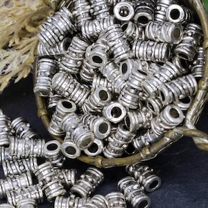 May include: A close-up of a silver metal bowl filled with silver metal beads. The beads are cylindrical with a textured surface and a small hole in the center. The bowl is sitting on a dark gray surface.