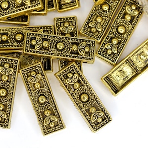 May include: A collection of rectangular gold-toned metal beads featuring an intricate embossed pattern with a central circular motif and symmetrical floral-like accents at each end, arranged in a scattered layout on a plain white surface.
