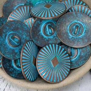 Puede incluir: Una colección de botones metálicos turquesa con un acabado envejecido. Los botones tienen un borde color cobre y presentan diseños en relieve, incluyendo líneas radiales y formas geométricas. Los botones están en un cuenco de color neutro.