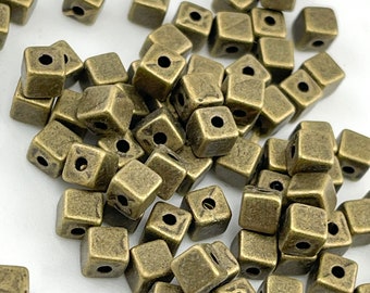 Könnte beinhalten: Eine Sammlung kleiner, quadratischer Perlen mit einem metallischen Bronzefinish. Jede Perle hat ein Loch in der Mitte, das zum Auffädeln geeignet ist. Die Perlen sind auf einer weißen Oberfläche verstreut und erzeugen ein strukturiertes Aussehen.