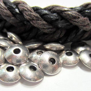 May include: Close-up of silver metal beads with a hole in the center. The beads are round and have a slightly domed shape. They are arranged in a pile on a white surface.