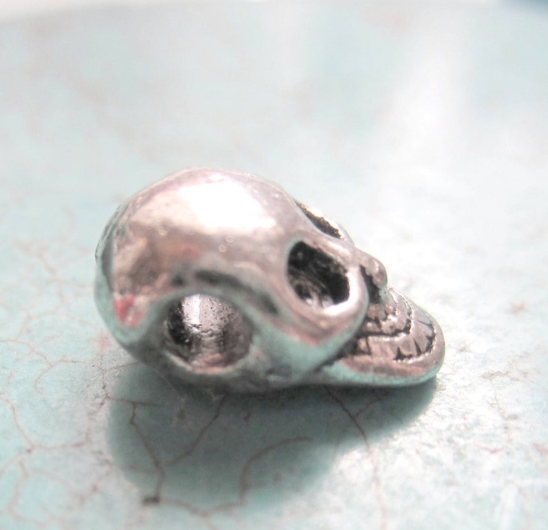 K&ouml;nnte beinhalten: Ein silberner Totenkopf-Anh&auml;nger mit einem Loch zum Einf&auml;deln. Der Totenkopf ist detailliert mit einem zahnigen Grinsen und Augenh&ouml;hlen.
