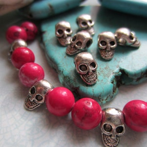 K&ouml;nnte beinhalten: Ein pinkfarbenes und silbernes Armband mit Totenkopfperlen. Das Armband besteht aus pinkfarbenen Perlen und silbernen Totenkopf-Anh&auml;ngern. Das Armband liegt auf einem t&uuml;rkisfarbenen Hintergrund.