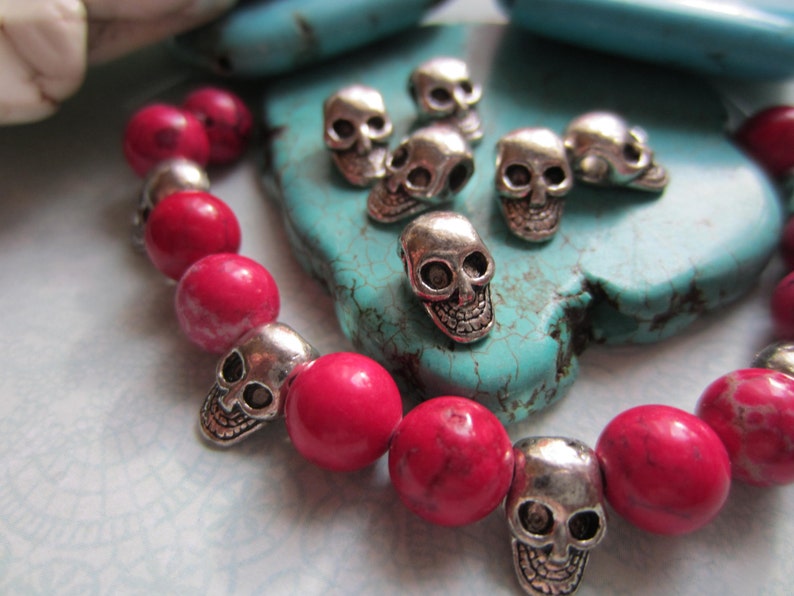 K&ouml;nnte beinhalten: Ein pinkfarbenes und silbernes Armband mit Totenkopfperlen. Das Armband besteht aus pinkfarbenen Perlen und silbernen Totenkopf-Anh&auml;ngern. Das Armband liegt auf einem t&uuml;rkisfarbenen Hintergrund.