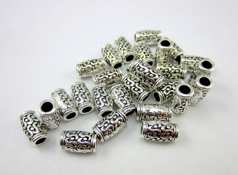 May include: A pile of silver-toned metal beads with an intricate, swirling design. The beads are cylindrical and have a smooth, polished finish.