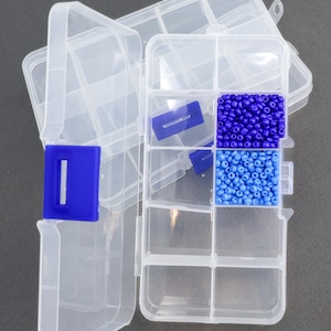 May include: Clear plastic craft organizer with a blue snap closure and multiple compartments. The organizer contains blue beads in two shades. The open lid is visible in the background.