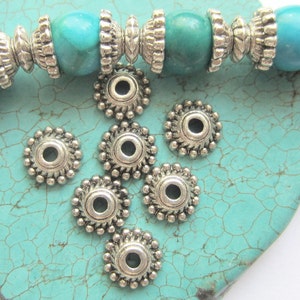 May include: Silver metal bead caps with a decorative design. The caps are round with a small hole in the center. They are arranged on a turquoise background.