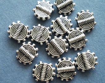 May include: A collection of silver-colored metal gear-shaped beads. Each bead features a spiral design within the gear and a horizontal bar across the center. The beads are scattered on a light blue surface.