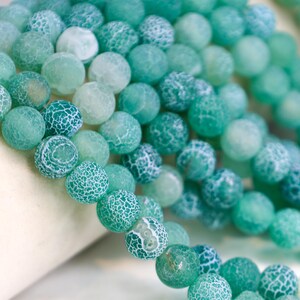 May include: Close-up of a collection of round, teal-colored beads with a cracked, white pattern. The beads are strung together, creating a textured and visually appealing design. The beads vary in shades of teal and green.