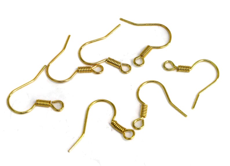 May include: Several gold-colored earring hooks are scattered on a white surface. Each hook features a curved design with a coiled spring detail near the top, and a straight, pointed end for insertion into the ear.