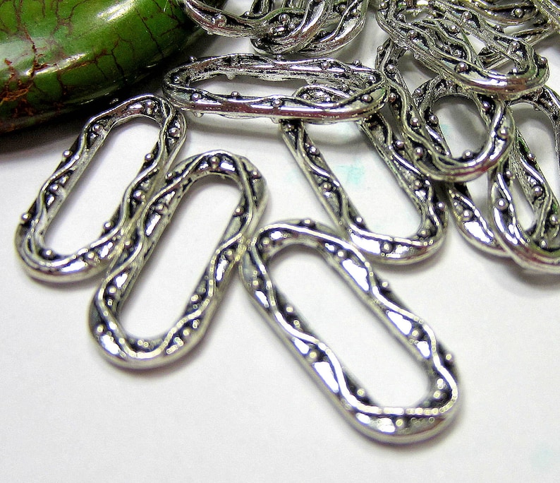 May include: Silver metal oval-shaped jump rings with a decorative design. The rings have a textured surface with small raised dots.