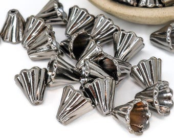 Op de afbeelding: Een verzameling zilverkleurige, kegelvormige metalen kralen met een gegroefd ontwerp. De kralen zijn verspreid op een wit oppervlak, met enkele gedeeltelijk zichtbaar in een kleine, gebroken witte kom. De kralen hebben een glanzende, metalen afwerking.