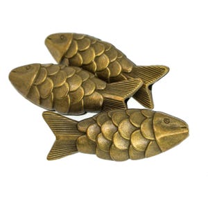 May include: Three metallic fish-shaped ornaments with an antique brass finish. Each piece features a detailed scale pattern, a defined head with a small eye, and a textured tail fin. The fish are arranged in a cluster, showcasing their curved bodies.