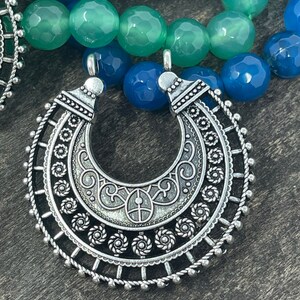 May include: A silver pendant with an intricate design, featuring a crescent moon shape with a circular border. The pendant is made of metal and has a detailed, ornate pattern.