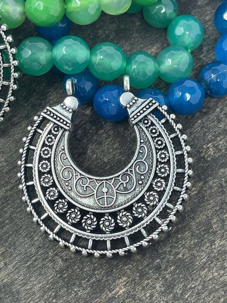 May include: A silver pendant with an intricate design, featuring a crescent moon shape with a circular border. The pendant is made of metal and has a detailed, ornate pattern.
