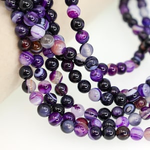 May include: A strand of round, polished beads in various shades of purple, violet, and deep black. The beads feature natural banded agate patterns with translucent and opaque sections, creating a smooth, glossy texture throughout the necklace.