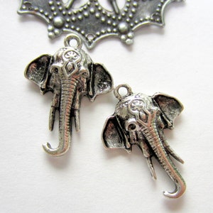 May include: Two silver-colored elephant head charms with detailed trunks and ears, and a decorative crown-like element. The charms have a loop for attaching to jewelry or other crafts. The background is white.