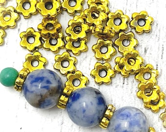 May include: Close-up of a bracelet with blue and white stone beads, gold-colored flower-shaped bead caps, and green beads. The beads are strung together, creating a decorative jewelry piece.