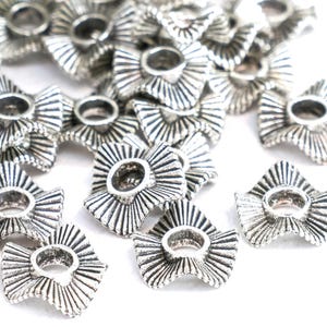 May include: A collection of silver-colored metal flower-shaped beads. Each bead has a central hole and a textured, fan-like design radiating outwards. The beads are scattered on a white surface.