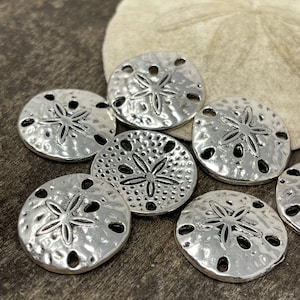 May include: A group of eight silver-toned metal sand dollar charms with a raised star design. The charms have a hammered texture and are arranged on a brown wooden surface.