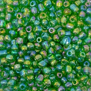 May include: A close-up shot of a collection of small, iridescent green beads. The beads have a shiny, reflective surface with hints of yellow and blue. The beads are cylindrical with a hole through the centre, suitable for stringing.