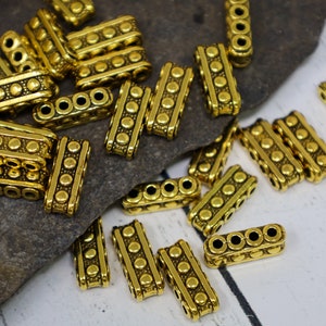 May include: Close-up of several gold-colored metal beads. Each rectangular bead has three holes and is decorated with a series of small, raised dots. The beads are scattered on a gray stone surface.