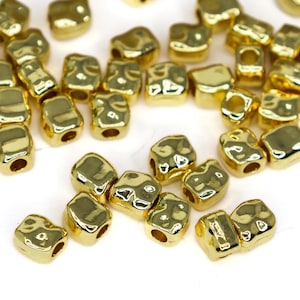 Puede incluir: Un conjunto de cuentas brillantes de color dorado con superficies irregulares y texturizadas. Cada cuenta tiene un agujero que la atraviesa, lo que sugiere que están diseñadas para ensartar. Las cuentas están dispersas sobre un fondo blanco.