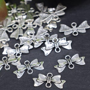 May include: A pile of silver-toned metal bow charms with two loops on each bow. The charms are scattered on a black surface.