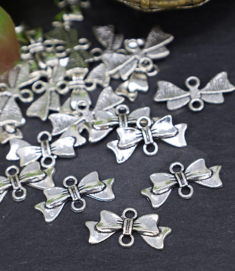 May include: A pile of silver-toned metal bow charms with two loops on each bow. The charms are scattered on a black surface.