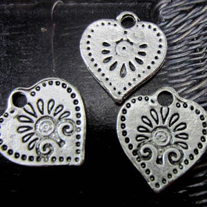 May include: Three silver heart-shaped charms with a decorative floral design. Each charm has a small loop at the top for attaching to jewelry or other crafts.