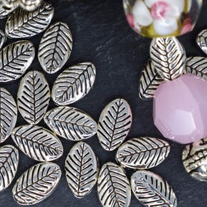 May include: A collection of silver metal leaf-shaped beads. The beads are oval-shaped and have a textured surface.