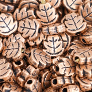 May include: Close-up of a pile of copper-colored leaf-shaped beads. Each bead has a detailed leaf vein design in a darker shade, with a hole for stringing. The beads are ideal for jewelry making and other craft projects.