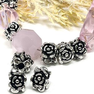 May include: A close-up of a bracelet featuring rose-shaped silver beads and pink gemstone beads. The bracelet has a mix of faceted and rounded beads, creating a delicate design. The silver beads add a touch of elegance to the piece.