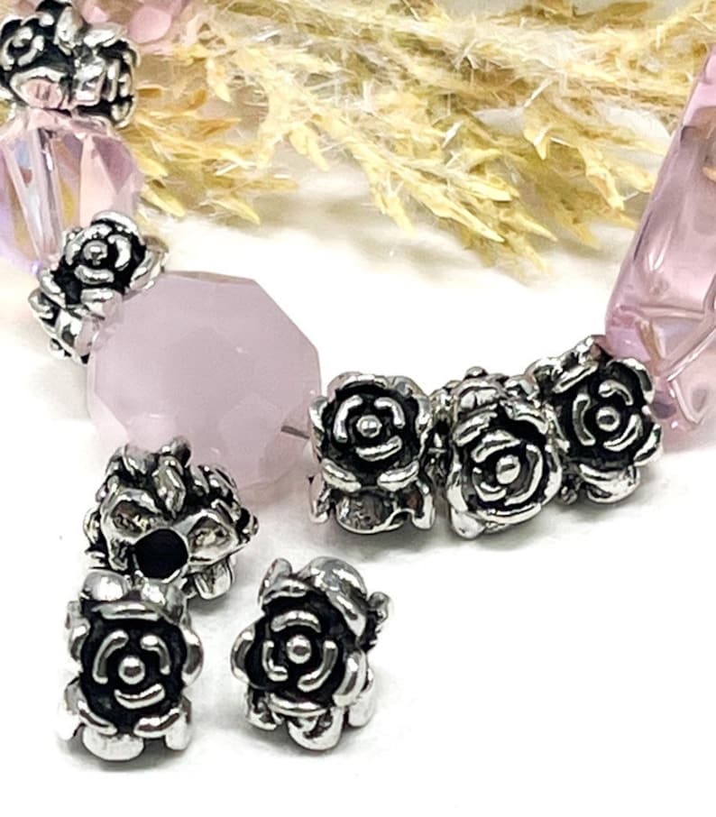 May include: A close-up of a bracelet featuring rose-shaped silver beads and pink gemstone beads. The bracelet has a mix of faceted and rounded beads, creating a delicate design. The silver beads add a touch of elegance to the piece.