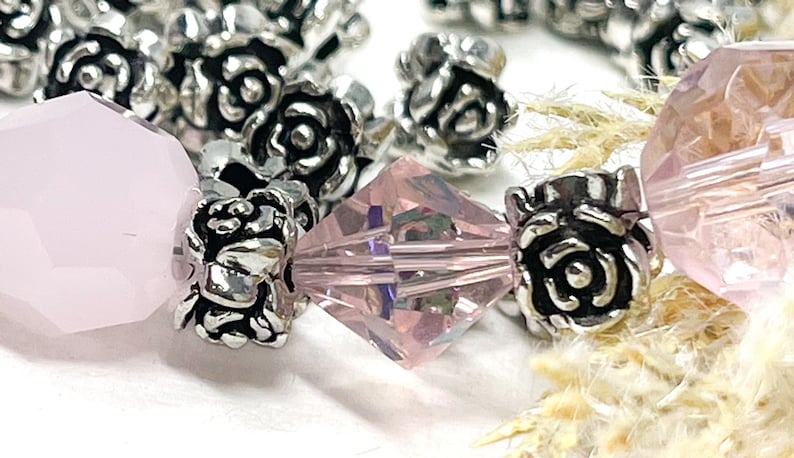 May include: Close-up of a bracelet with pink faceted beads and silver rose-shaped spacers. The beads are translucent, and the silver spacers add detail. The bracelet is set against a soft, neutral background.
