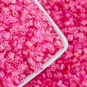May include: Close-up of a white square bowl filled with translucent pink seed beads. The beads are cylindrical with a small red dot in the center of each. The background is filled with more of the same pink beads.