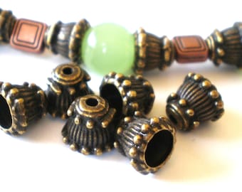 May include: A collection of bell-shaped metal beads with an antique bronze finish and a ribbed texture, featuring small raised dots along the base. In the background, a partially visible string of beads includes copper-colored squares and a light green bead.