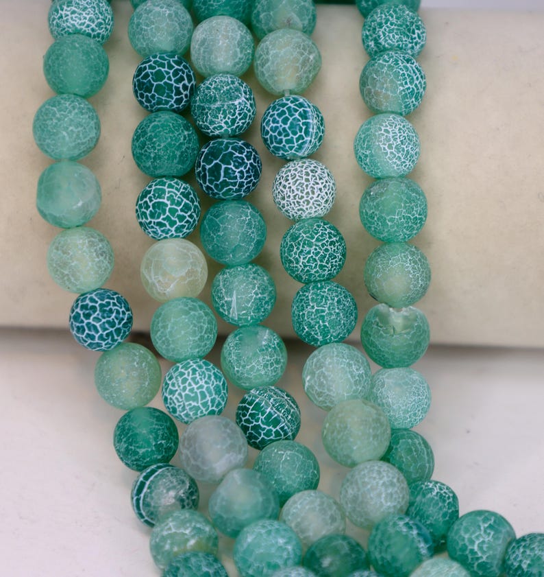 May include: Strands of round, green beads with a cracked, web-like pattern. The beads vary in shades of green, from light to dark, and are strung together. These beads are suitable for jewelry making.