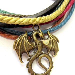 May include: A multi-colored braided leather bracelet with a bronze dragon charm. The bracelet is made of brown leather, beige rope, red rope, blue rope, and green rope.