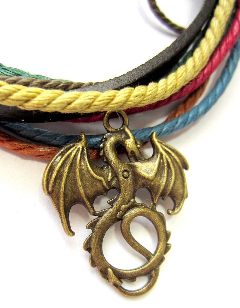May include: A multi-colored braided leather bracelet with a bronze dragon charm. The bracelet is made of brown leather, beige rope, red rope, blue rope, and green rope.